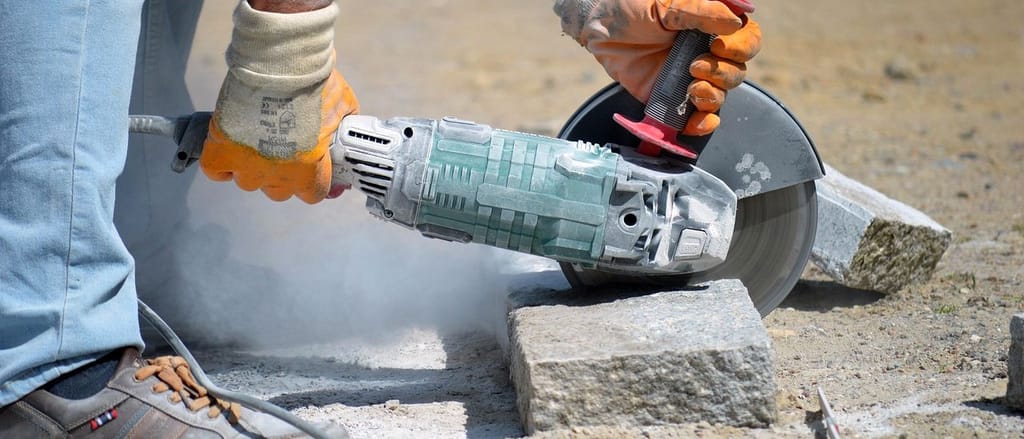 Hände in Arbeitshandschuhen trennen einen Granitstein mit dem Winkelschleifer, Staub steigt auf.